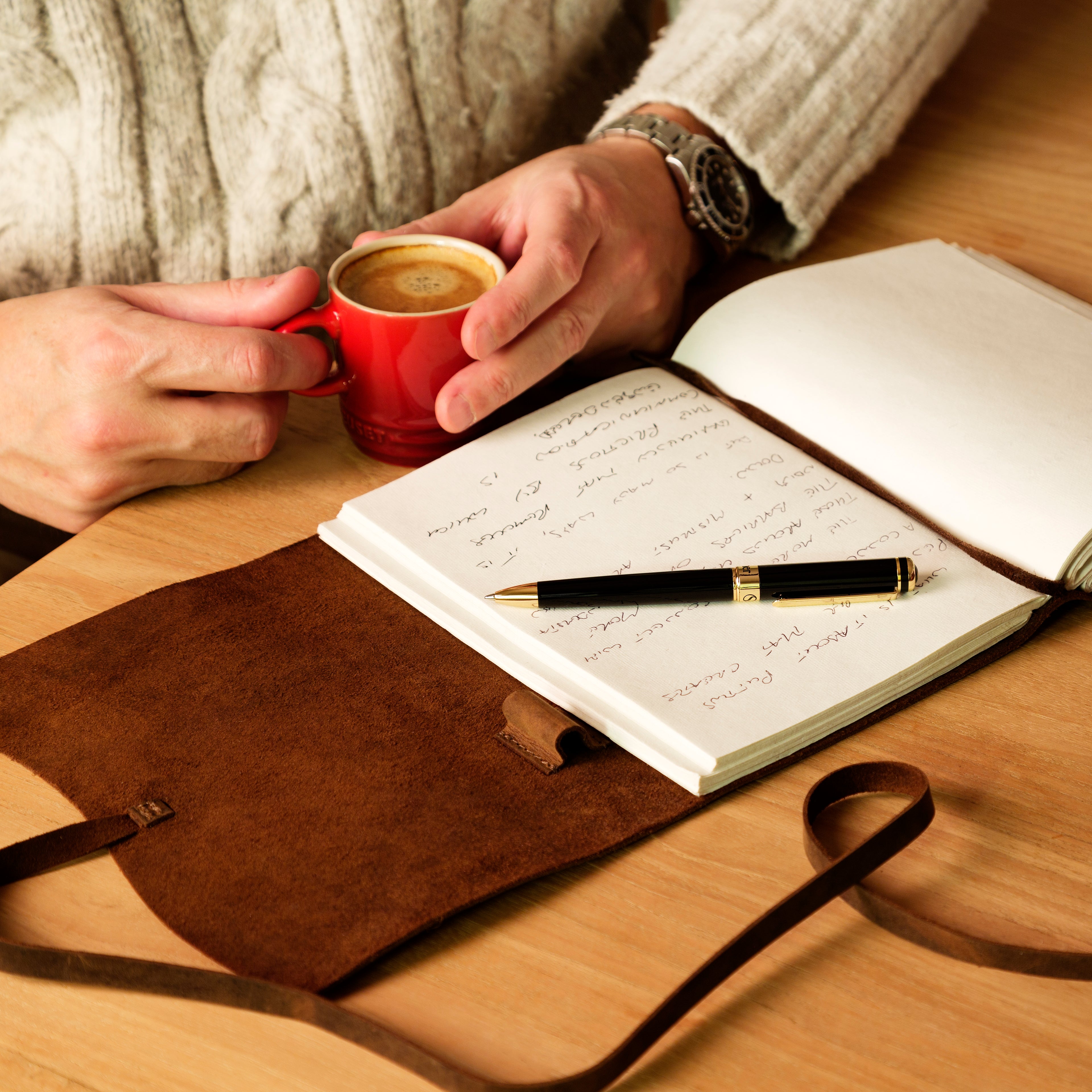Person holding a red mug with a cup of coffee next to an open notebook and pen on a wooden table.