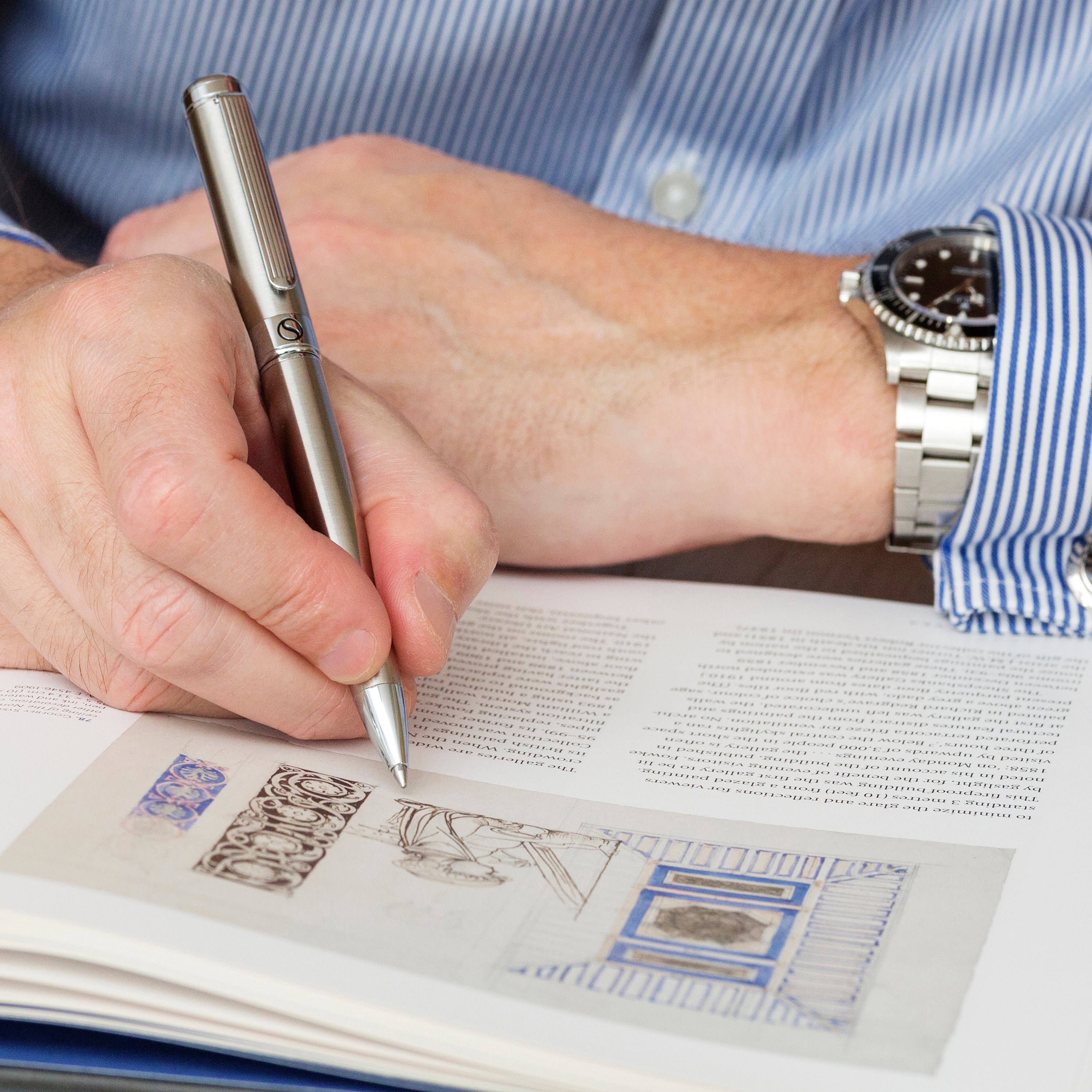 Person holding a pen over an open book with detailed illustrations.