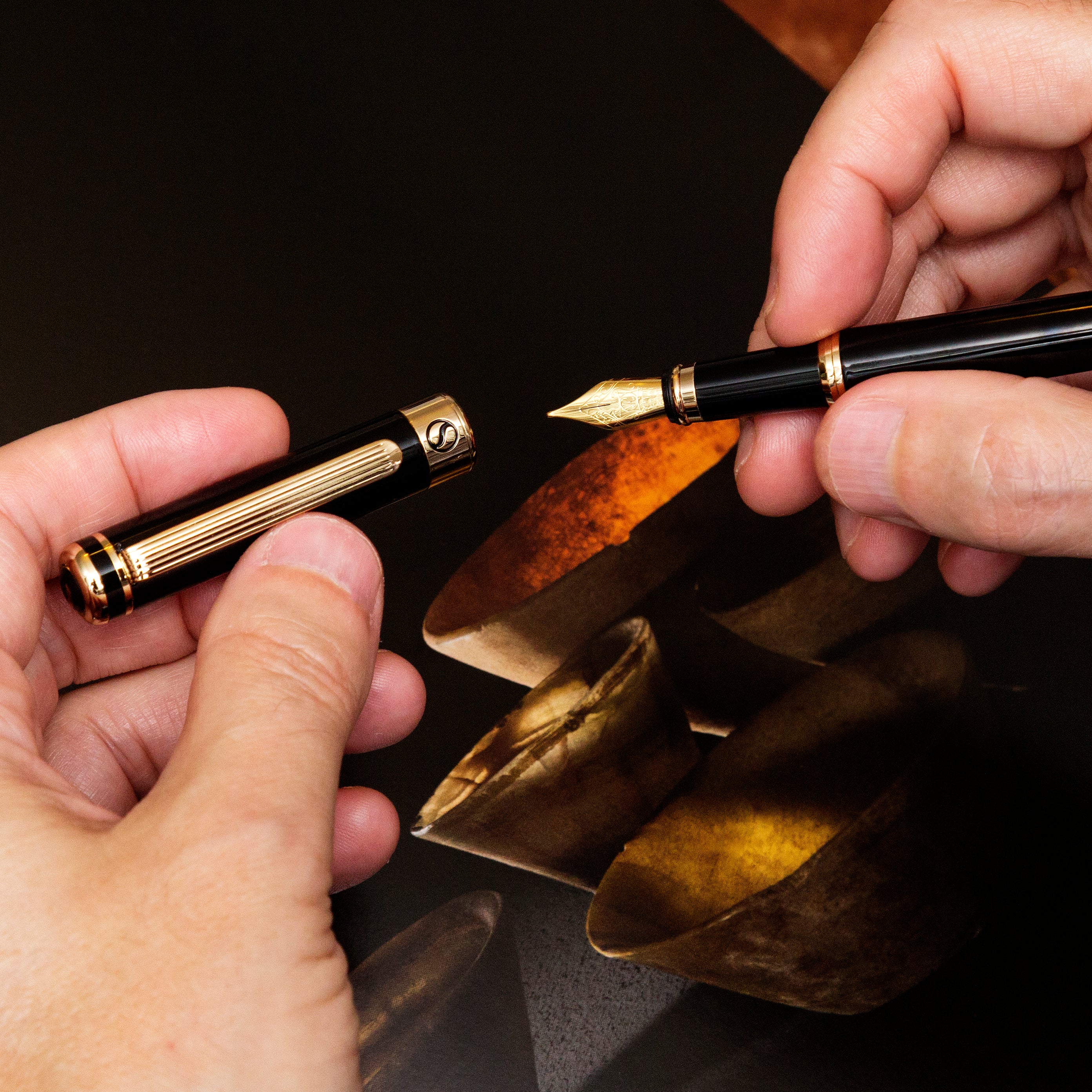 Two hands holding a black and gold pen with a close-up of the nib against a dark background.
