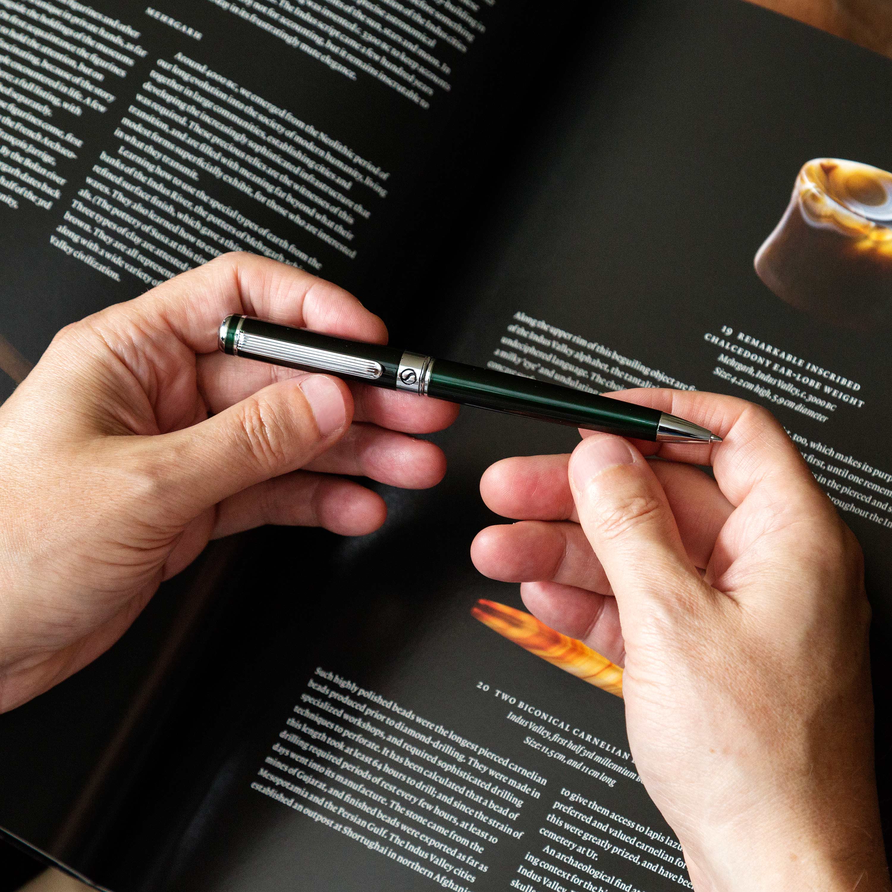 Person holding a green pen over a magazine with a blurred coffee cup image.
